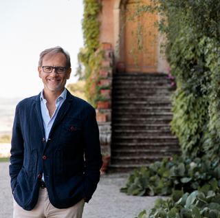 Giampiero Bertolini, CEO of Biondi‑Santi, smiles while standing next to an ivy‑covered wall. He wears a light blue shirt, a navy blazer, and glasses.