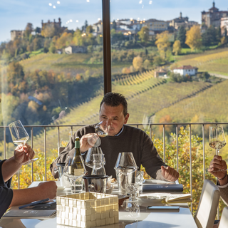 Cogno Wines winemaker Valter Fissore sitting at the head of a table, smelling a glass of wine, with his vineyards visible behind him.
