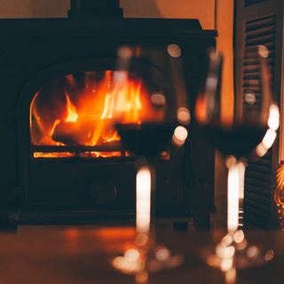 Two red wine glasses in the foreground, blurred, with a roaring fireplace in focus behind them.
