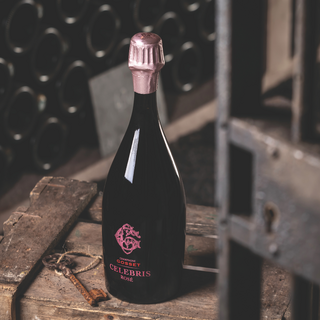 A bottle of Champagne Gosset Celebris Rosé with pink foil and pink lettering sits on a wooden surface in a wine cellar, surrounded by old wooden boards and a vintage key tied with twine, with rows of bottles blurred in the background.