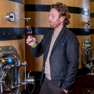 Federico Radi, head winemaker of Biondi-Santi, stands in a cellar wearing a dark gray blazer, holding up a glass of Sangiovese and examining it closely.