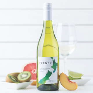 A bottle of Frenzy Marlborough Sauvignon Blanc stands centered on a light surface, with sliced kiwi, grapefruit, peach, and lime arranged around it; a partially filled wine glass sits in the background against a clean white backdrop.
