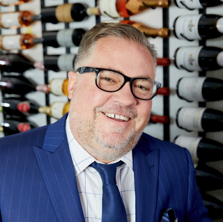 A screenshot of Chris Pedersen in the Wilson Daniels wine cellar. He is smiling, and wearing a blue suit and glasses.