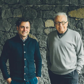 Photo of Alberto Graci and Angelo Gaja standing side by side in front of a gray stone wall. Alberto is on the left, wearing a buttoned-up cardigan, and Angelo is on the right dressed in gray.