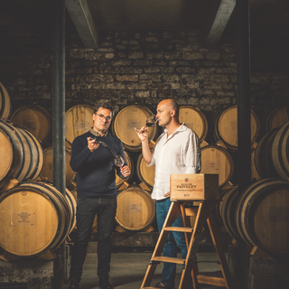 Domaine Faiveley Winemaker Jérôme Flous and Proprietor Erwan Faiveley standing together in the cellar, surrounded by rows of aging barrels and stone walls.