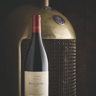 Bottle of Te Mata Estate Bullnose Syrah in front of a large copper-colored metal object with a textured grille pattern, photographed in dramatic low lighting against a dark background.