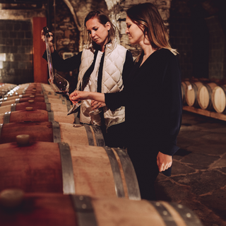 Sisters Karoline and Julia Walch with red wine together in a wine cellar surrounded by barrels.