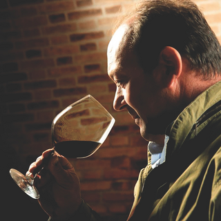 Romano Dal Forno in side profile, holding a glass of red wine near his nose, wearing a green jacket.