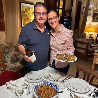 Fabio Sireci and Melissa Muller of Feudo Montoni stand beside a dining table set for a meal. Fabio holds a bottle of Feudo Montoni wine, while Melissa holds a platter of food.