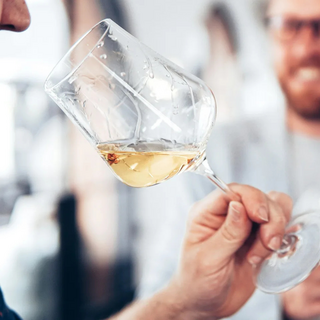 Close-up of a hand raising a glass of white wine to a person's nose for smelling.