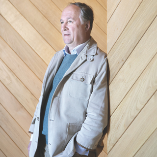 Portrait of Brice de La Morandiere standing against a light wood-paneled wall, wearing a beige jacket over a blue sweater and white collared shirt.