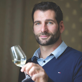 Olivier Bailly of Domaine Billaud-Simon pictured holding a glass of white wine. Olivier Bailly is wearing a light blue button-down shirt beneath a dark blue V-neck sweater and has short dark hair and blue eyes.