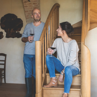 Erwan and Eve Faiveley of Domaine Faiveley hold glasses of red wine while posed on a wooden staircase, with Erwan standing beside the banister and Eve seated on the steps in a warmly lit interior.