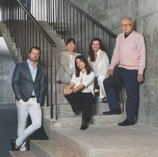 The Gaja family poses on a stone staircase, with fifth-generation Giovanni Gaja standing on the left in a dark suit jacket and light trousers, fourth-generation Angelo Gaja standing on the right in a pink sweater, and the rest of the family seated together on the steps between them.