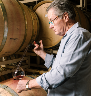 Winemaker Vance Rose of Harper's Rest draws red wine from an oak barrel into a tasting glass while standing among rows of aging barrels in the winery cellar.