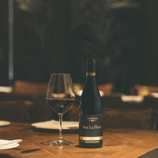 A bottle of Familia Torres Mas La Plana red wine displayed alongside a wineglass on a wooden table in a dimly lit restaurant setting.