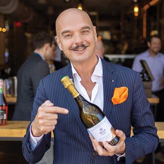 21st generation family member Gianluca Bisol smiles and points at one of his wine bottles. He is wearing a blue suit with an orange pocket square.