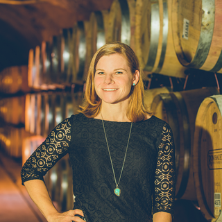A headshot of Danielle Ball in front of boxes of wine. She is wearing a brown plaid blazer and a chunky brown necklace.