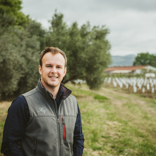 A photo of Guillaume Boudet, winemaker at Napa Valley’s Hyde de Villaine, outdoors. He is wearing winery branded zip-up vest and smiling at the camera.