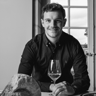 A black and white photo of Domaine Laroche Winemaker Romain Chevrolat sitting at a table, smiling warmly while posing with a glass of wine in hand.