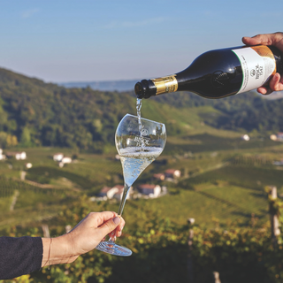 Against the dramatic, steep hills of Valdobbiadene, a hand on the right pours Bisol1542 Prosecco Superiore into a glass held by another hand on the left. The two hands frame the photo, capturing a moment of sharing amidst a stunning vineyard backdrop.