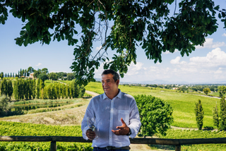Marco Caprai stands outdoors on the Arnaldo Caprai estate, wearing a light blue button-down shirt and holding a glass of wine as he speaks, surrounded by the vineyard landscape.