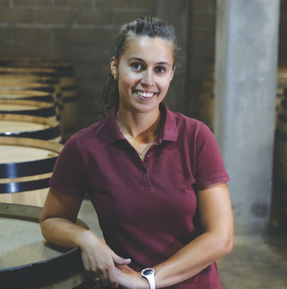 Juliette Alips, winemaker at Domaine Les Monts Fournois, stands in front of wine barrels wearing a purple short-sleeve polo with her hair tied up.